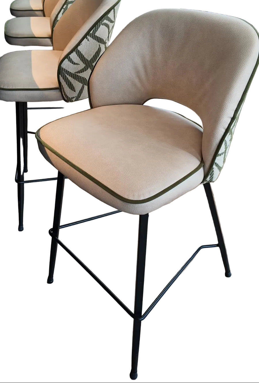 A modern bar stool with a curved beige upholstered seat and backrest, accented with dark green piping and patterned green-and-white side bolsters, set on a light wood floor.