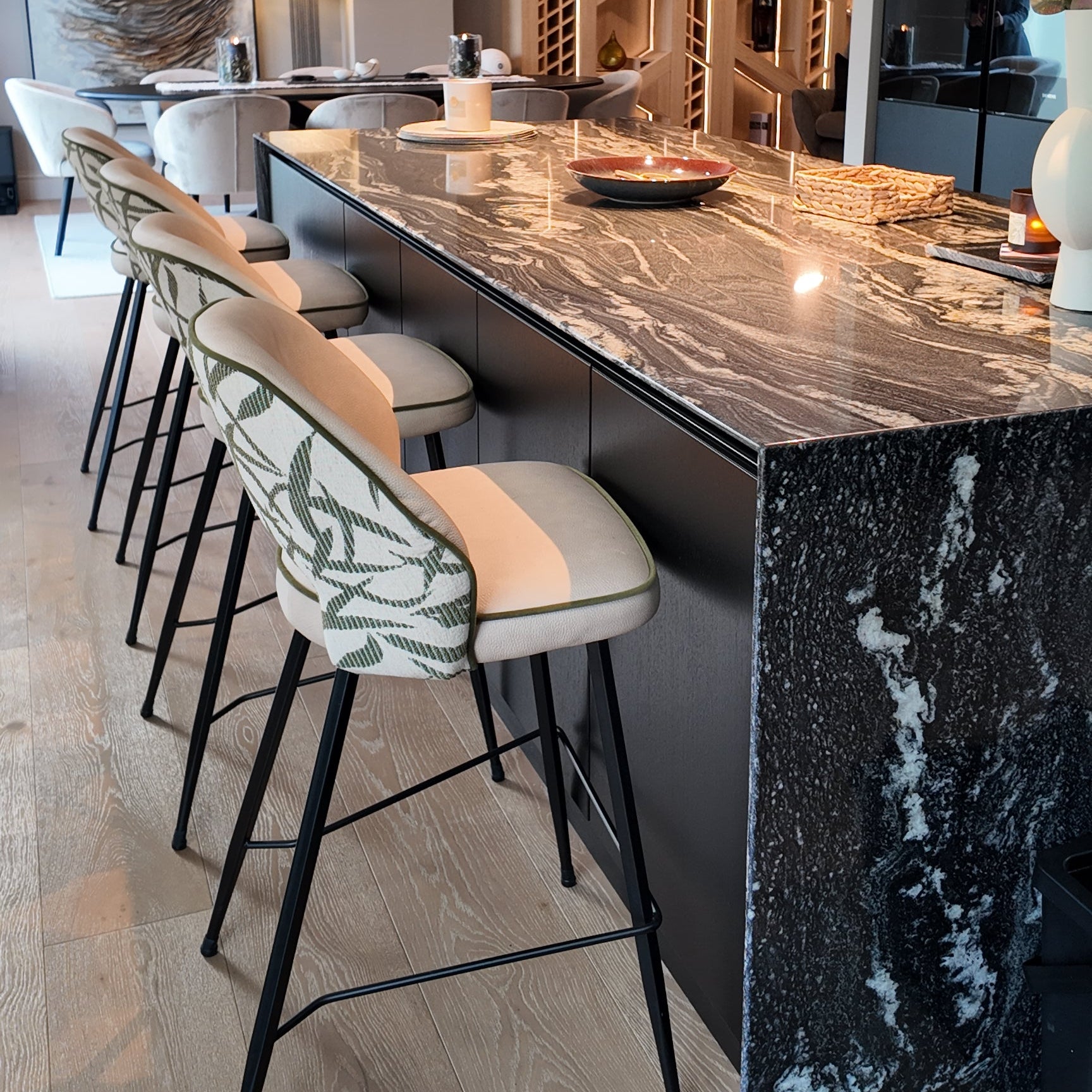 A modern kitchen island with a black and white marble countertop and matching marble backsplash, paired with four upholstered bar stools featuring cream cushions and green leaf-patterned fabric, set in a contemporary interior.